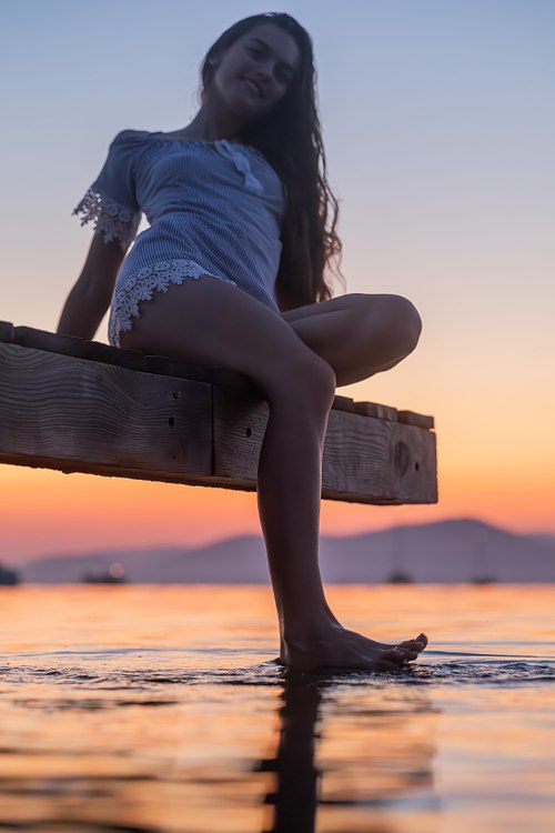 Femme assise sur un ponton photographiée en contre-plongée avec la mer et le ciel du soir