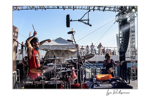 Horizontal color image of Mike Portnoy performing at the drum kit with drumstick raised on an outdoor stage while another musician plays nearby