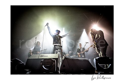 Horizontal color image of The Tragically Hip performing live on stage, with Gord Downie in a glittering silver suit at center with arms raised high while band members play under bright concert spotlights