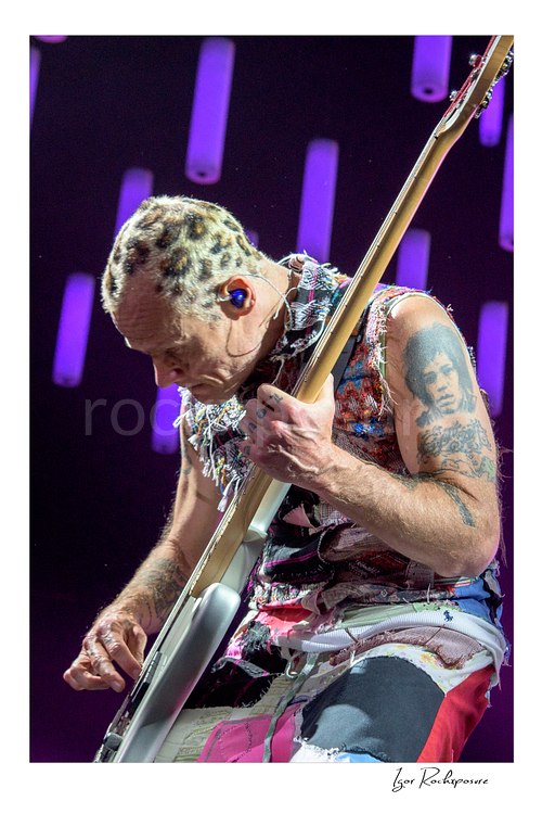 Flea bends over his bass with the neck angled high, patterned shaved hair and tattooed arms visible beneath glowing purple LED tubes in close-up.