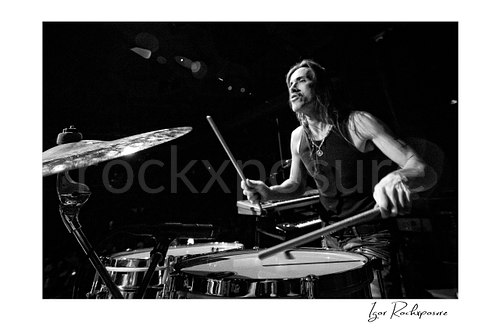 Horizontal black and white image of Nuno Bettencourt in an extreme close-up on stage playing drums during a live performance