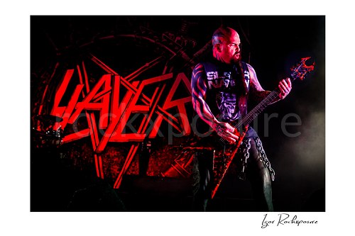 Horizontal color image of Kerry King of Slayer playing guitar onstage under intense red and blue concert lighting with the Slayer logo behind him