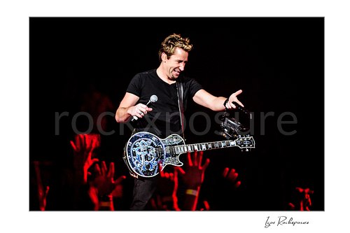 Horizontal color image of Chad Kroeger of Nickelback holding a microphone and guitar while reaching toward the crowd under red concert light