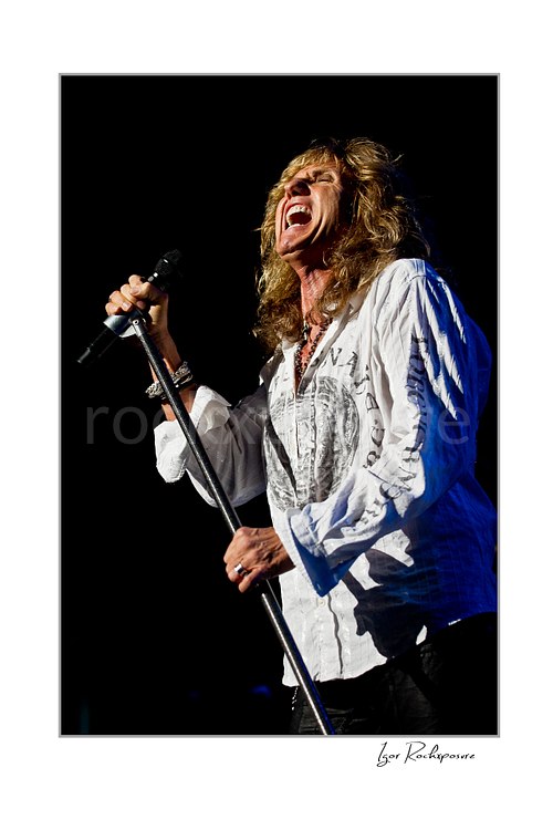 Vertical color image of David Coverdale of Whitesnake performing live on stage, singing into a microphone while holding a mic stand against a dark background