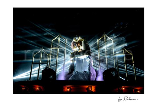 Horizontal color image of Maria Brink of In This Moment seated in a dramatic live performance pose on a raised platform, surrounded by bright white beams, stage smoke, and glowing frame props