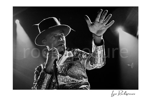 Horizontal black and white image of Gord Downie of The Tragically Hip holding a microphone and extending one hand forward, wearing a glittering stage jacket and hat under dramatic backlight