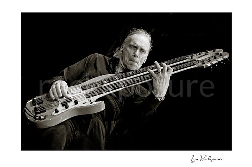Horizontal black and white image of Billy Sheehan playing his double-neck bass in a tight low-angle live performance shot