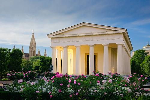 Weißer Theseustempel im Wiener Volksgarten, umrahmt von blühenden rosa und weißen Rosenbüschen, im Hintergrund ist der Turm des Wiener Rathauses zu sehen.