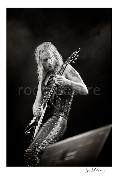 Vertical black and white image of Richie Faulkner of Judas Priest performing live on stage, playing flying V guitar against a dark background