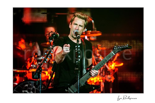 Horizontal color image of Chad Kroeger of Nickelback singing into a microphone and pointing toward the crowd while holding a guitar under vivid red concert light