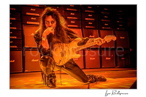 Horizontal color image of Yngwie Malmsteen kneeling on stage with a white guitar under intense warm orange lighting, framed by a wall of Marshall amplifiers behind him
