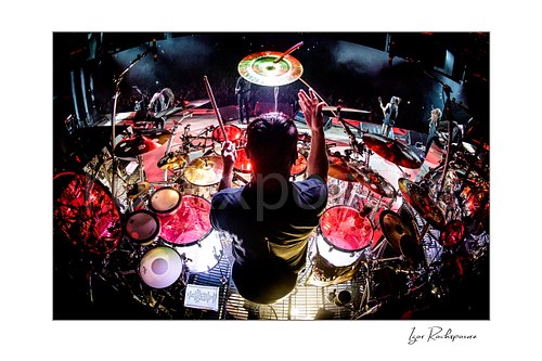 Horizontal color image of Korn drummer Ray Luzier seen from behind at the drums with one drumstick raised above the stage and crowd