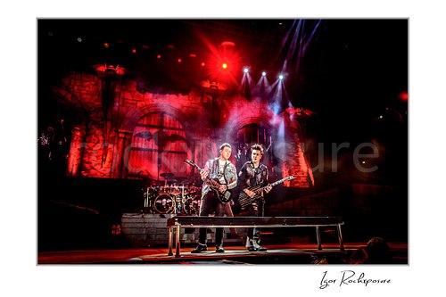 Horizontal color image of Synyster Gates and Johnny Christ of Avenged Sevenfold performing on stage together under red light with the drum kit behind them