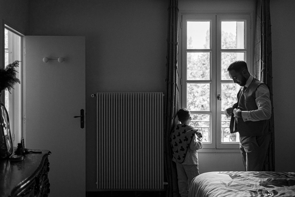 Un moment p&egrave;re-fils touchant, avec un jeune gar&ccedil;on regardant par la fen&ecirc;tre et le mari&eacute; en costume faisant son n&oelig;ud de cravate, symbolisant l'importance de la famille lors du mariage c&eacute;l&eacute;br&eacute; dans une prairie la&iuml;que pr&egrave;s de N&icirc;mes