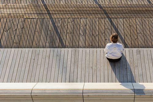 Person sitter på ett trädäck i kvällssol med långa skuggor och grafiska linjer – redaktionellt miljöporträtt i Stockholm.