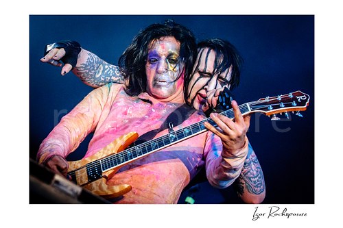 Horizontal color image of Marilyn Manson performing with a guitarist onstage in theatrical makeup under blue concert light