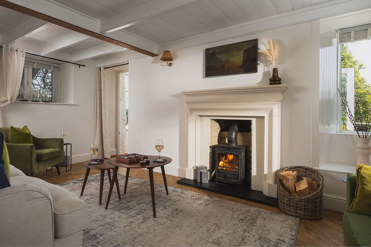 A cozy living room with a lit wood-burning stove, two small tables with wine glasses, a sofa, armchairs, and a window letting in natural light.