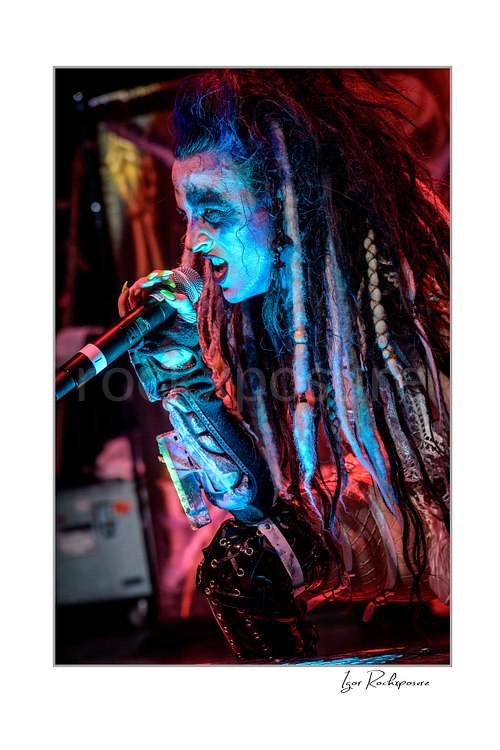 Vertical color image of Calico Cooper of Beasto Blanco singing into a microphone in a fierce live performance close-up, with long dreadlocks, dramatic makeup, and vivid blue-red stage light