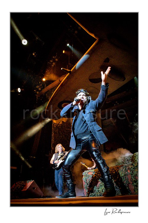 Bruce Dickinson of Iron Maiden performing live on stage, singing into a microphone with one arm raised beneath the band’s airplane stage prop.