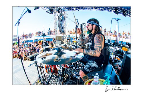 Horizontal color image of Mike Portnoy playing drums on an outdoor stage in a wide fisheye concert scene with the crowd behind him
