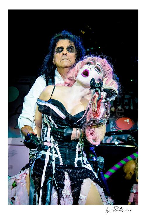 Vertical color image of Alice Cooper and Sheryl Cooper performing live on stage in theatrical makeup and costume with pink hair, black corset, and a dramatic horror prop