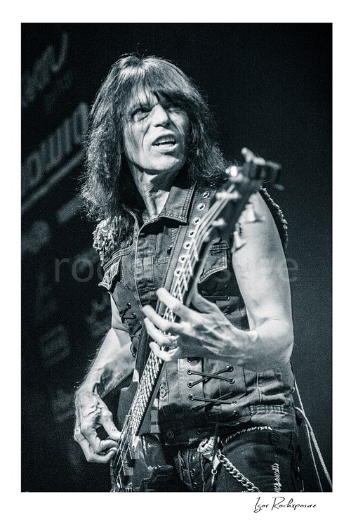 Vertical black and white image of Rudy Sarzo performing live with bass guitar in a close-up stage portrait