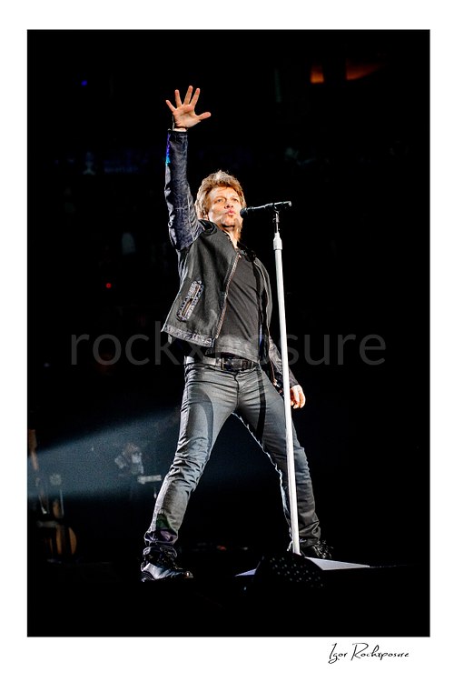Vertical color image of Jon Bon Jovi on stage beside a microphone stand, raising one hand high in a dramatic live performance pose