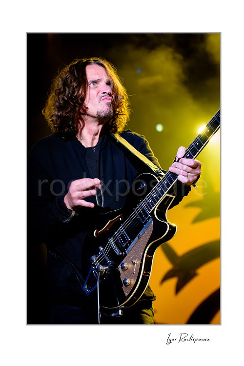 Vertical color image of Chris Cornell performing live on stage, holding his guitar upright under dramatic blue and red concert lighting