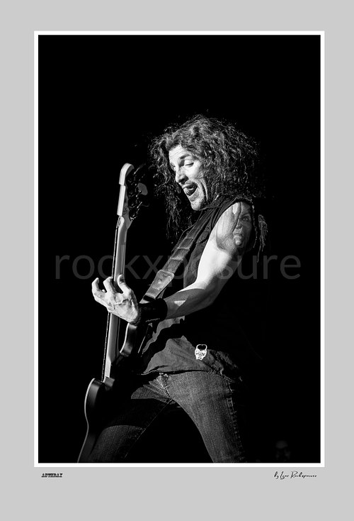 Vertical black and white image of Frank Bello of Anthrax performing live on stage, playing bass guitar against a dark background