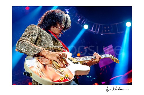 Horizontal color image of Joe Perry of Aerosmith performing live on stage with a custom painted guitar, glitter jacket, and blue concert spotlights