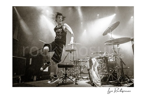 Horizontal black and white image of Zoltan Chaney performing beside his drum kit under bright stage lights