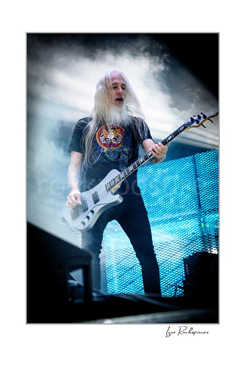 Vertical color image of John Campbell of Lamb of God playing bass on stage surrounded by thick smoke and bright backlight