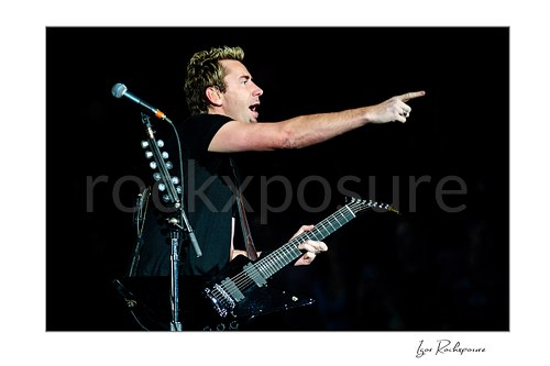 Horizontal color image of Chad Kroeger of Nickelback holding a guitar and pointing across the stage beside a microphone stand during a live performance