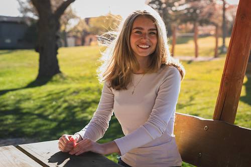Eine junge Frau mit langen blonden Haaren sitzt lächelnd an einem Holztisch in einem sonnigen Park bei Gegenlicht.