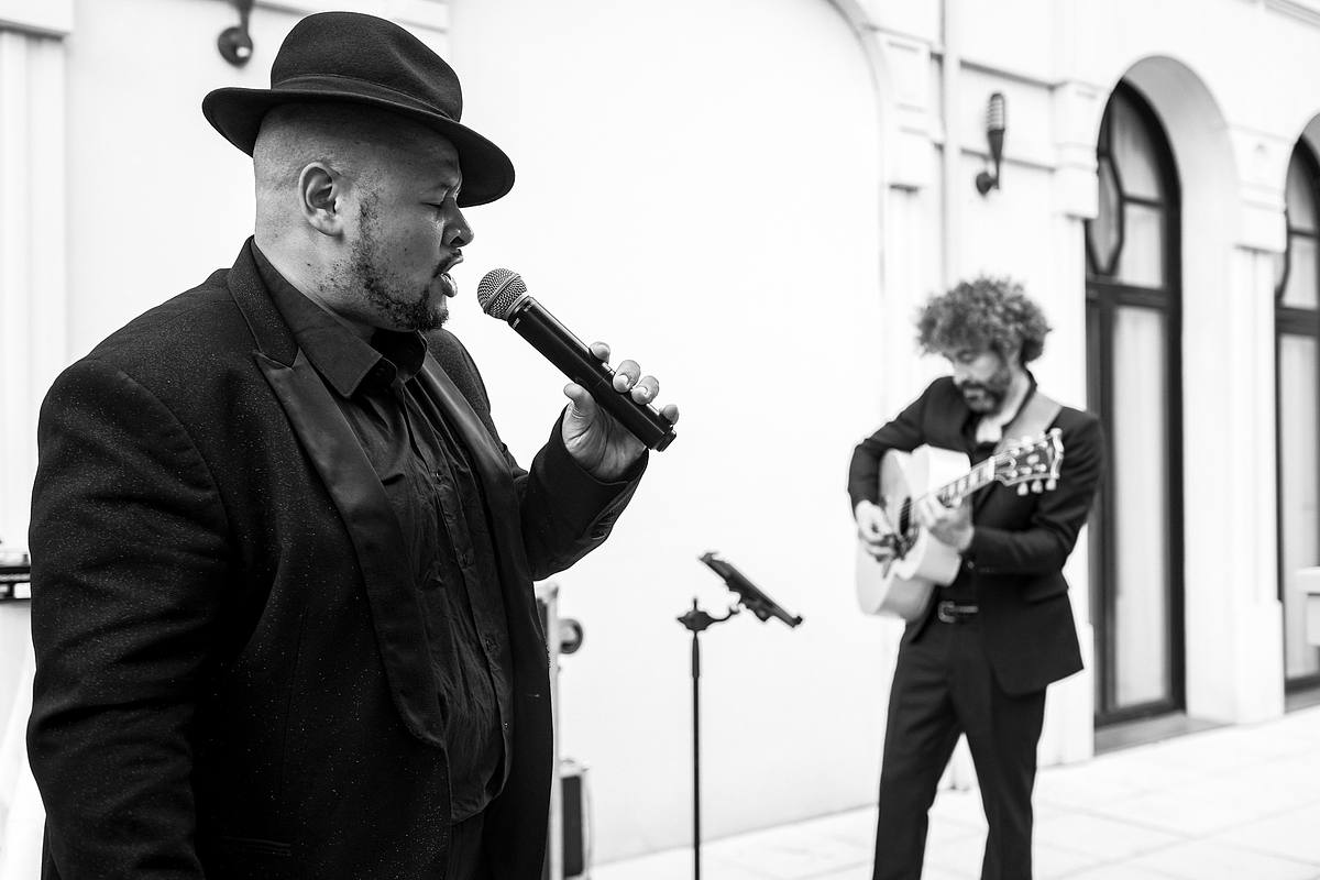 Sous un chapeau tendance, un chanteur du 'Groupe Love' livre une interpr&eacute;tation passionn&eacute;e accompagn&eacute; par un guitariste, devant un public attentif en noir et blanc, saisi par Sebastien CLAVEL Photographe mariage Lyon