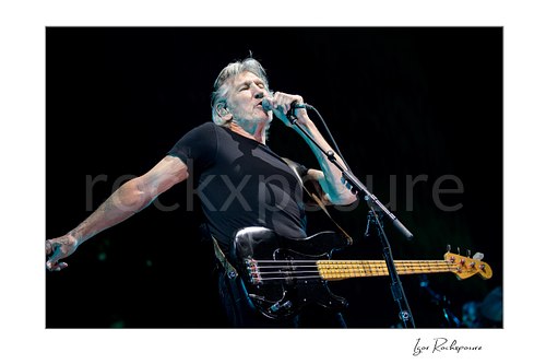 Roger Waters sings into a microphone with one arm extended sideways, a black bass hanging across his waist against a dark concert stage.