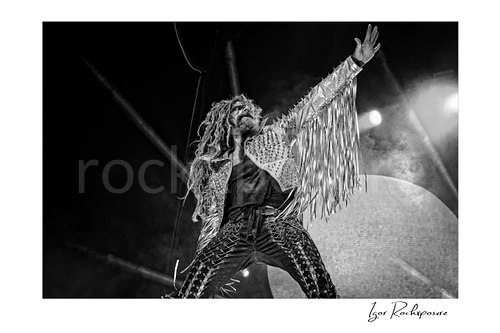 Horizontal black and white image of Rob Zombie performing live on stage in a fringed jacket with raised arms, handheld microphone, and a giant horned stage figure behind him