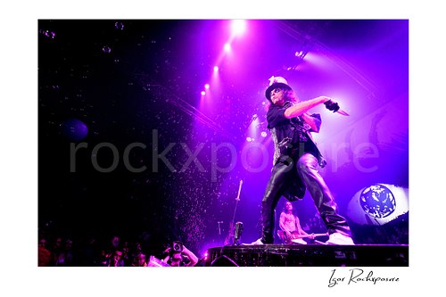 Horizontal color image of Alice Cooper performing live on stage under purple lighting with a raised knife, leather outfit, and falling bubbles