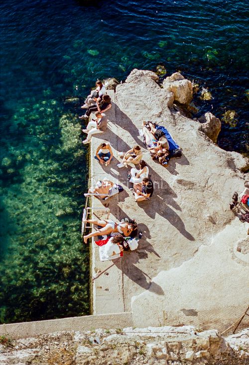 Photographie argentique de personnes installées au bord de l’eau dans une calanque à Marseille, scène estivale méditerranéenne par Dimitri Eteni