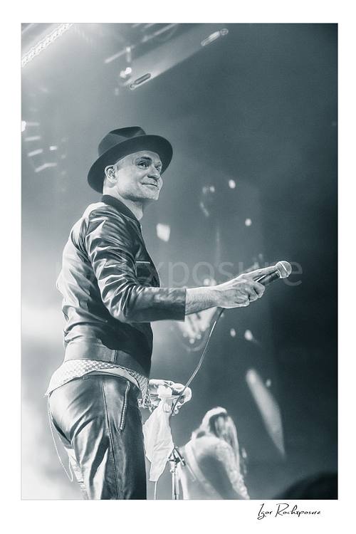 Vertical black and white image of Gord Downie of The Tragically Hip holding a microphone on stage, wearing a hat and leather jacket with bright concert haze and a blurred band member behind him