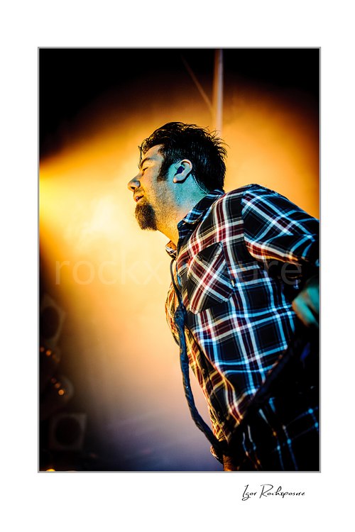 Vertical color image of Chino Moreno of Deftones performing live on stage in a plaid shirt with warm orange backlight and dramatic side-profile pose