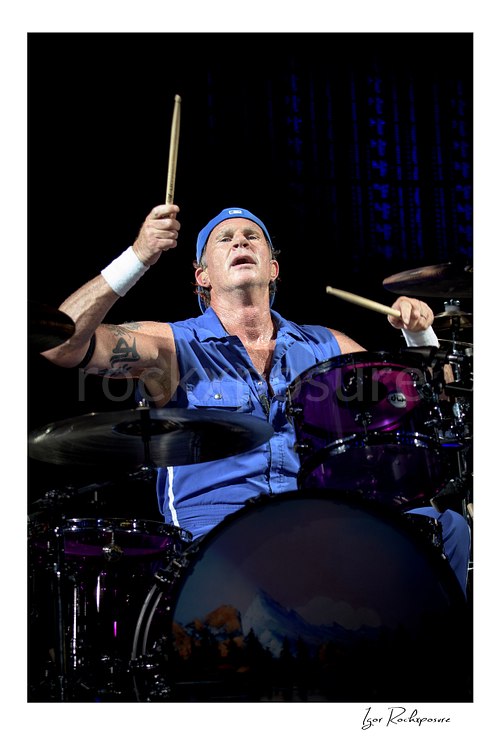 Chad Smith looks upward with both drumsticks lifted above his purple kit, wearing a blue cap and sleeveless shirt against a dark concert background.
