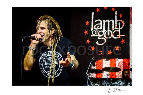 Horizontal color image of Randy Blythe of Lamb of God singing into a handheld microphone on stage with dreadlocks and the band backdrop behind him