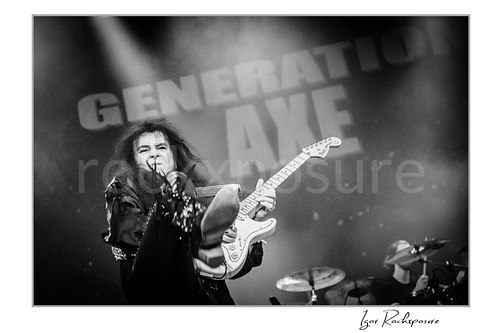 Horizontal black and white image of Yngwie Malmsteen performing live with a white guitar and delivering a dramatic high kick, framed by stage haze with the drummer visible behind him