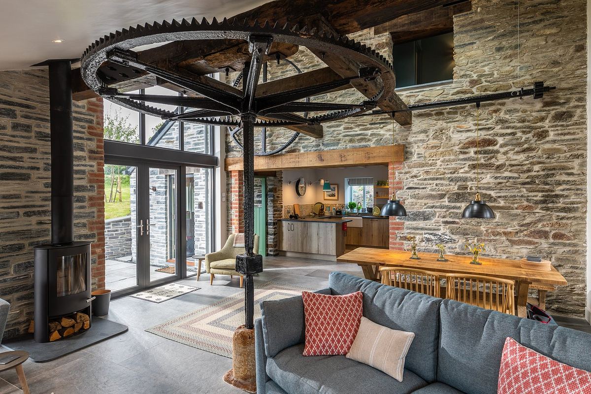 A living room with stone walls and a large metal wheel on the ceiling, the remnants of the building's previous use in mining. It features a grey sofa, dining table, and modern kitchen. Captured beautifully by an architectural photographer in Cornwall, natural light pours through the expansive windows and doors.
