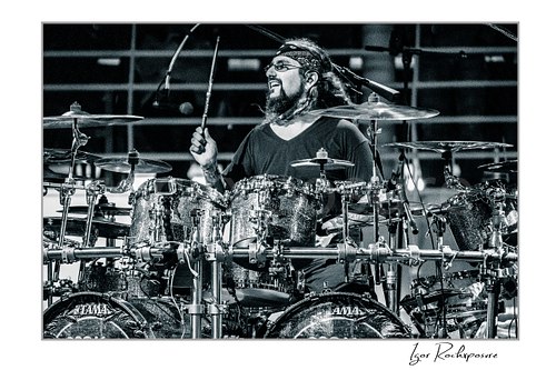 Horizontal black and white image of Mike Portnoy smiling at the drum kit during a live performance