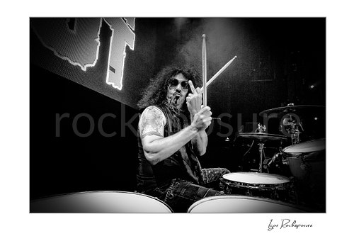 Horizontal black and white image of Frankie Banali at the drum kit holding drumsticks and raising his middle finger in a tight live performance shot