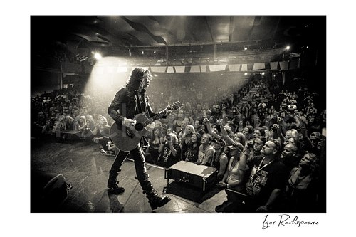 Horizontal black and white image of Tom Keifer performing live on stage with an acoustic guitar, walking toward the crowd under a bright spotlight with fans reaching forward