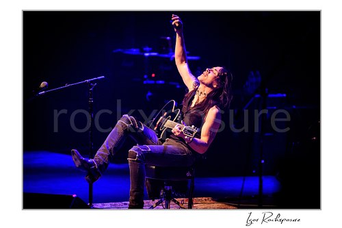 Horizontal color image of Nuno Bettencourt seated on stage with a guitar, one arm raised, under deep blue lighting