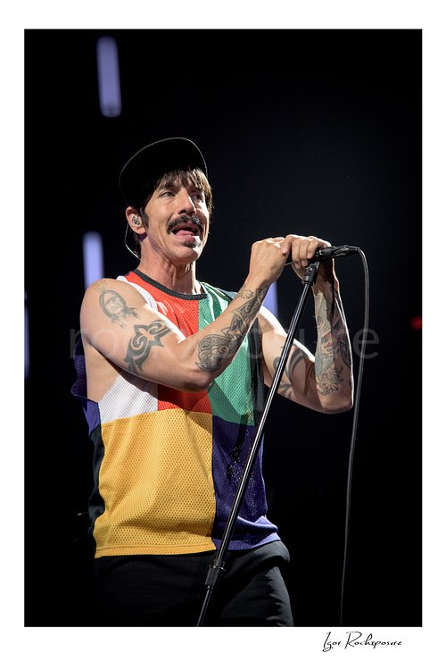 Anthony Kiedis holds a stand microphone with both hands, singing in a multicolor sleeveless top and black cap with tattooed arms against a dark stage.